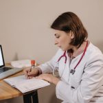 Médica escreve uma receita em um consultório, ao lado de um laptop.