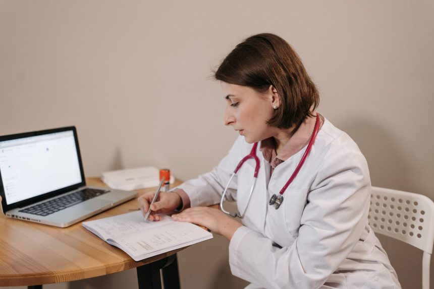 Médica escreve uma receita em um consultório, ao lado de um laptop.