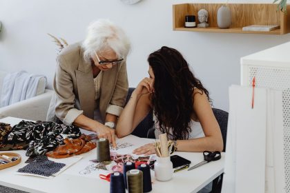 Mulher idosa orienta jovem em uma mesa de trabalho de um estúdio de moda.
